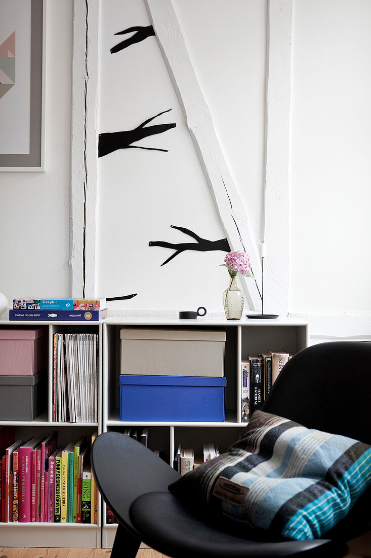 Schwarzer Stuhl mit Kissen vor Regal mit Büchern, darüber stilisierter Ast als Wanddekoration
