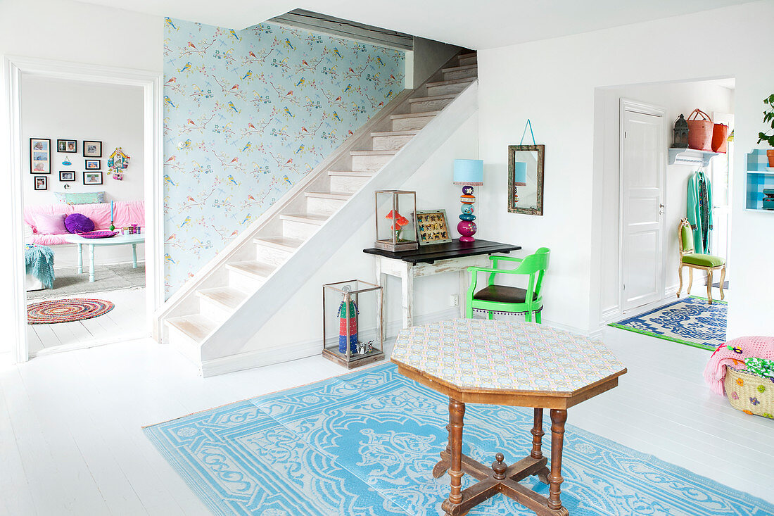 Achteckiger Holztisch auf blau-weißem Teppich im Vorraum mit weißem Holzboden, im Hintergrund Treppenaufgang