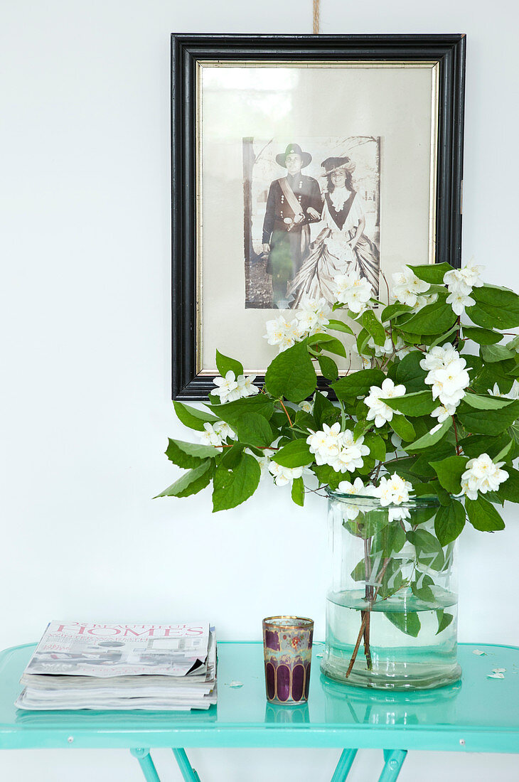 Zweig mit weißen Blüten in Glasvase auf türkisfarbenem Tisch, altes, gerahmtes Foto an der Wand