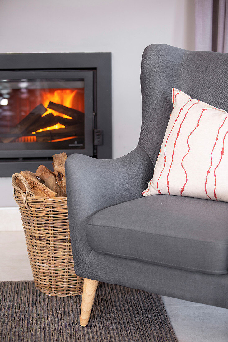 Grey armchair and basket of wood in front of fire in glass-fronted fireplace
