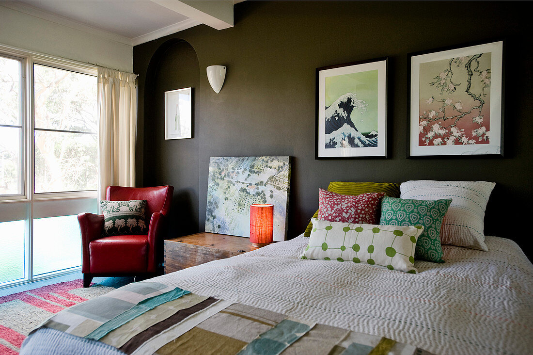 Bett mit Kissen und Stoffmuster, Nachtkasten und roter Ledersessel, Bilder an dunkler Wand im Schlafzimmer