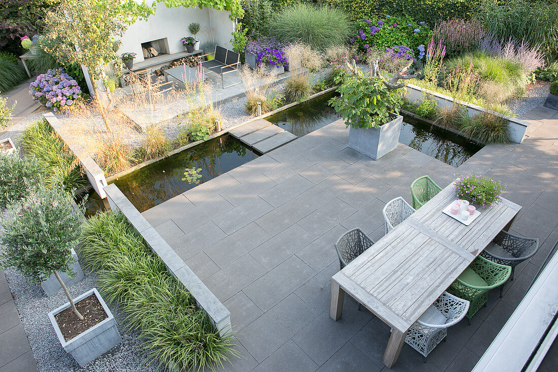 Holztisch und Designerstühle auf Terrasse mit Betonfliesen, umgeben von L-förmigem Teich