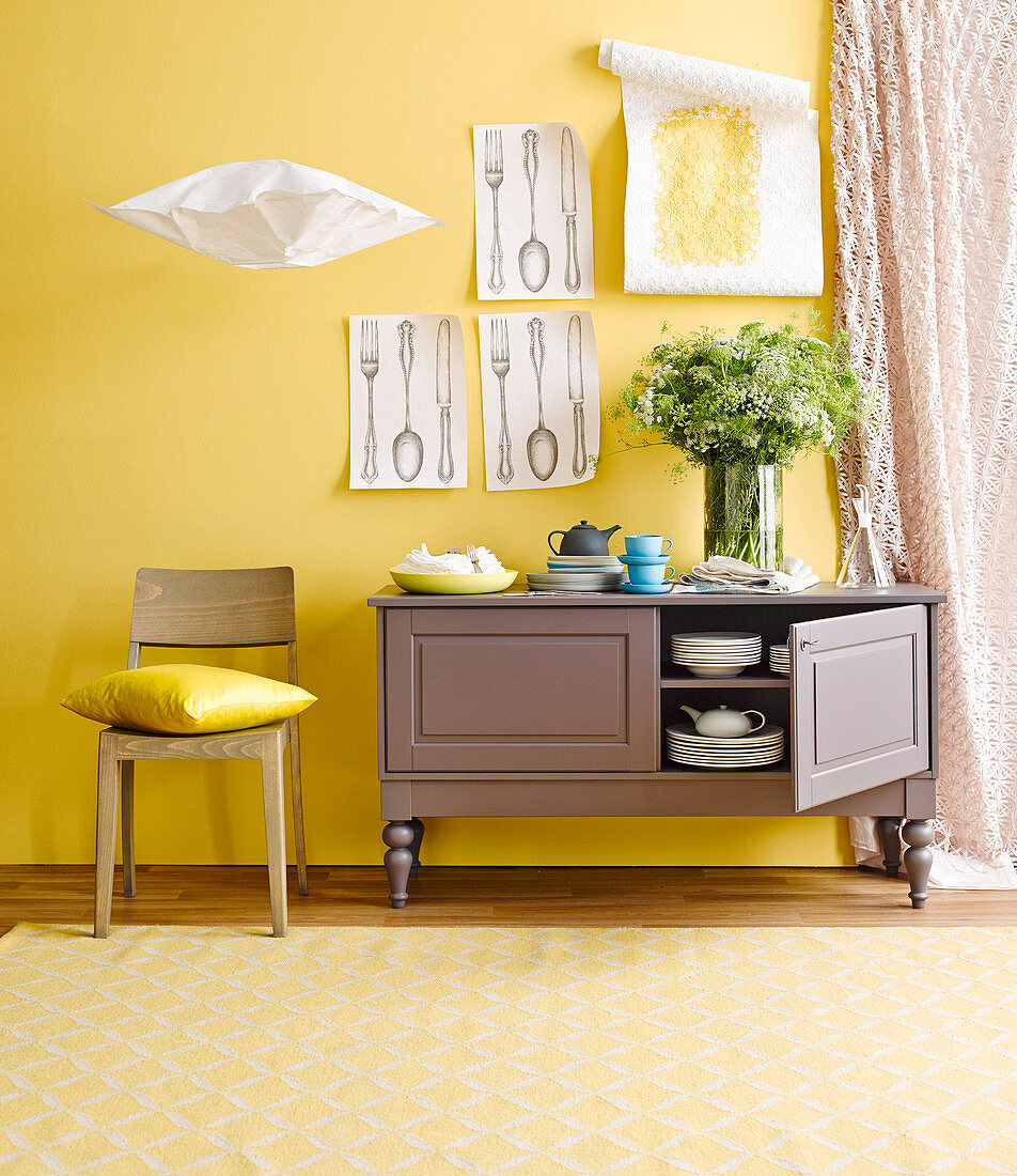Open sideboard with dishes in front of yellow wall with drawings