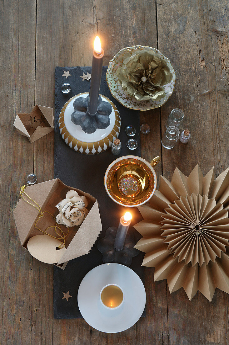 Weihnachtliche Tischdeko mit goldenem Porzellan, Papierblumen, Stern und grauen Kerzen auf Schieferplatte