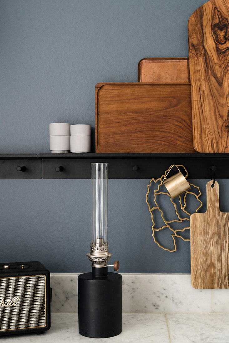 Holzbretter und Tabletts auf schwarzer Ablage, Petroleumlampe auf Marmor-Arbeitsplatte in der Küche mit blau-grauer Wand