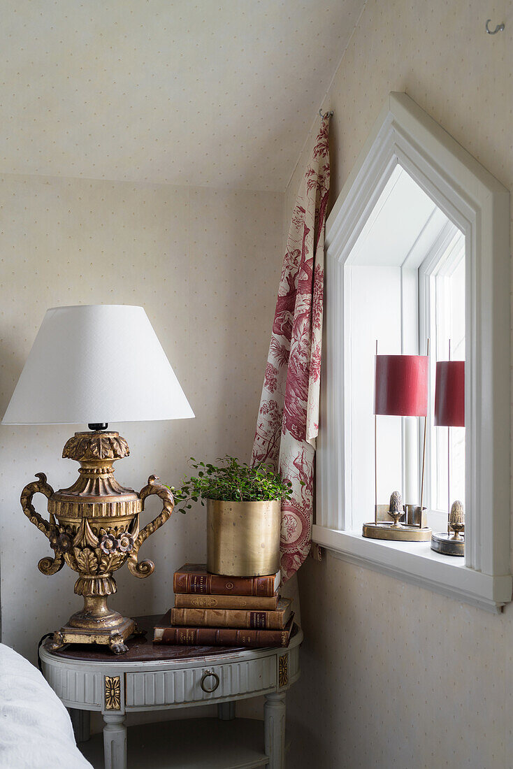 Antique table lamp on bedside table next to window with triangular arch in renovated Swedish house