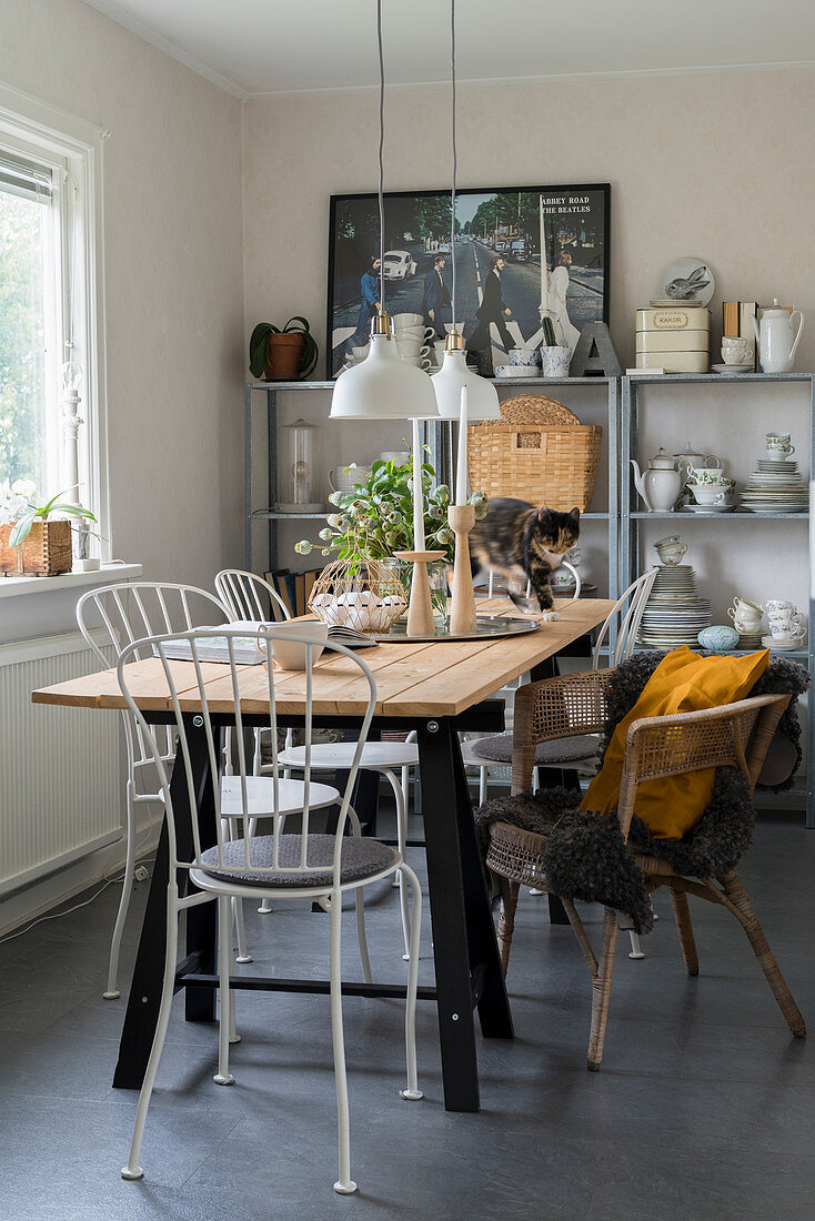 Custom-made table, white chairs and wicker armchair in front of crockery on shelves in dining area