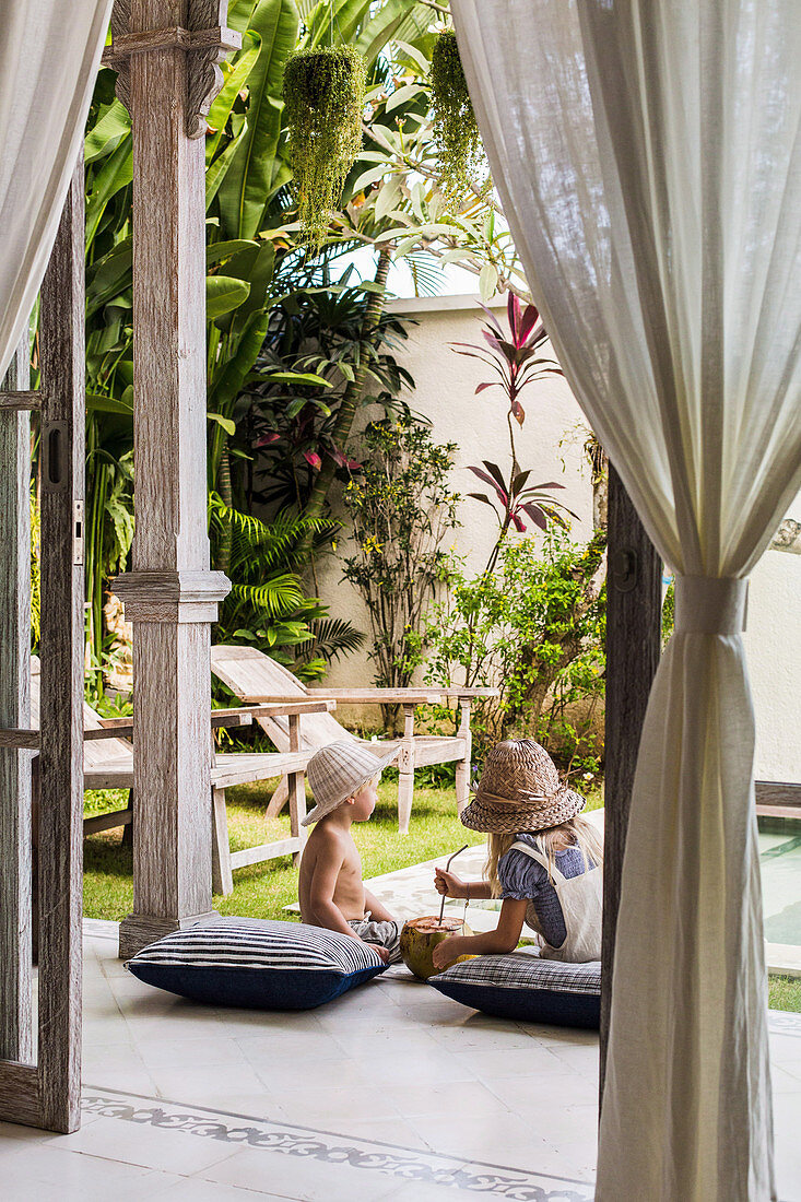 Zwei Kinder sitzen mit Kissen auf der Terrasse im exotischen Garten