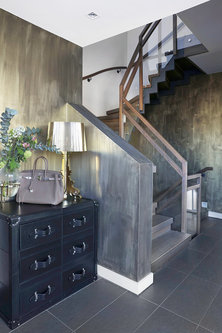 Table lamp on chest of drawers next to staircase in hallway in dark shades