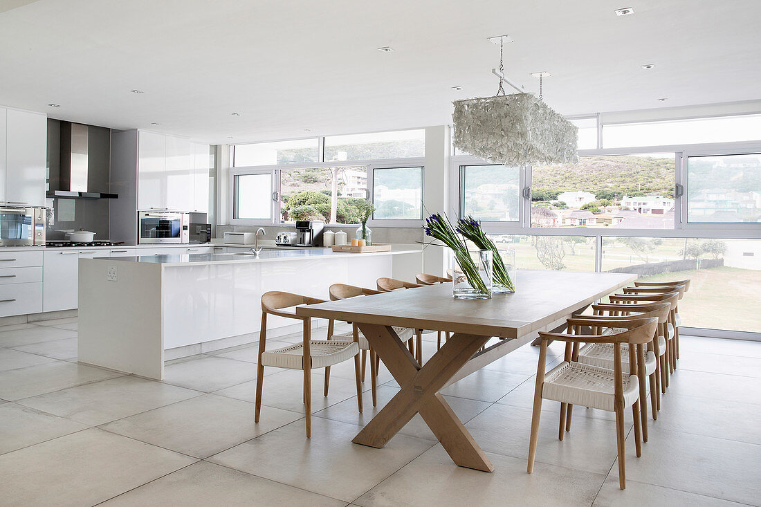 Long dining table in front of modern open-plan kitchen in interior with glass wall