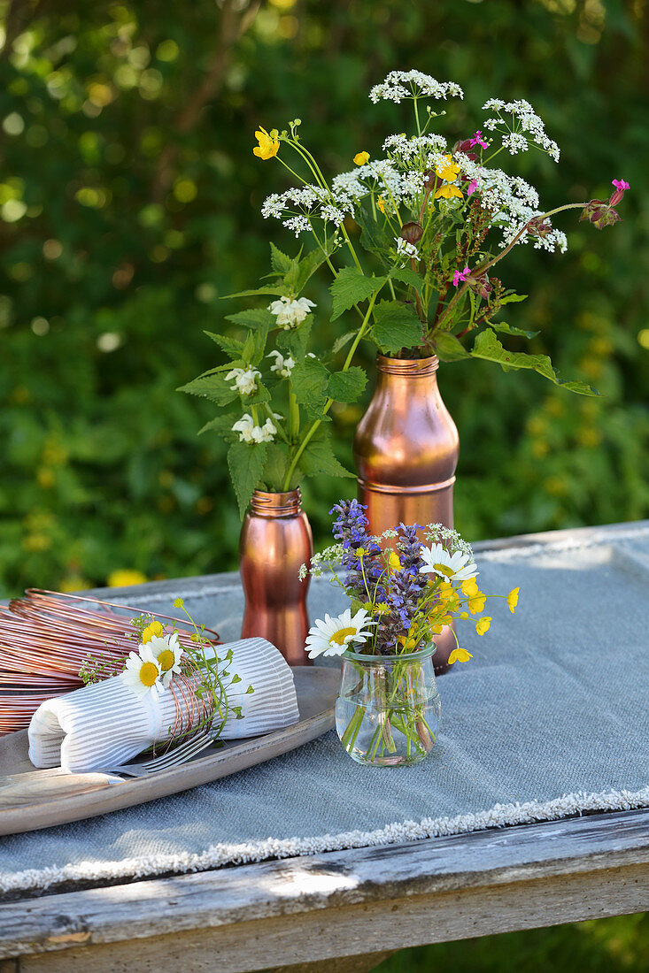 Sommerliche Wiesenblumensträuße in kupferfarbenen Gefäßen auf Tisch im Freien