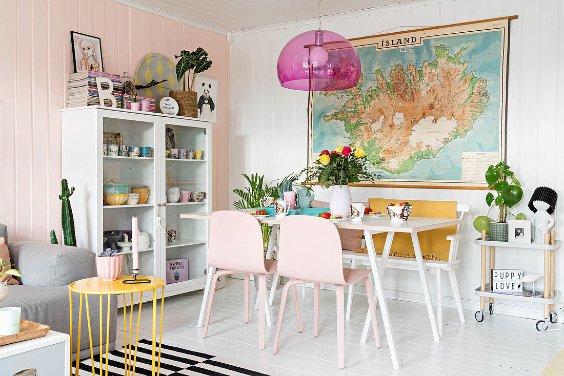 Essbereich mit weißem Tisch, Bank und rosa Stühlen dahinter Landkarte als Wanddekoration