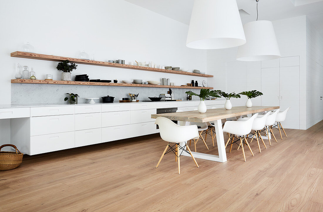Long white kitchen counter below wooden shelves and dining table with shell chairs