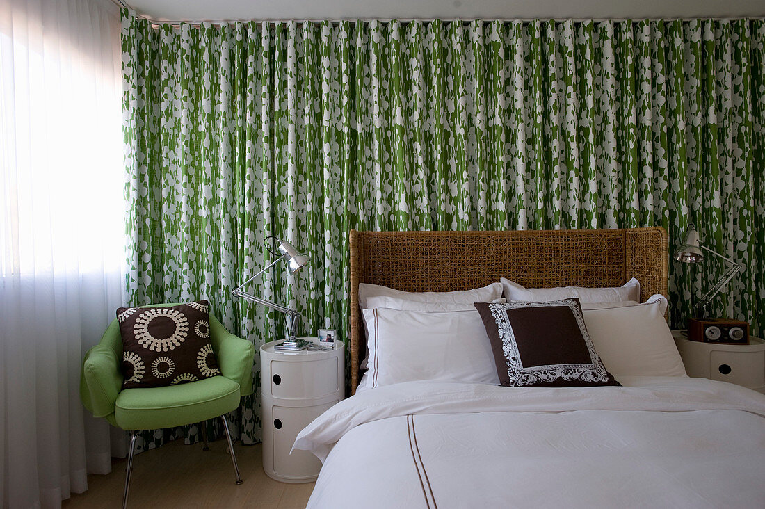 Doppelbett mit Rattan-Kopfteil, , Nachttisch und grüner Stuhl vor grün-weißem Vorhang im Schlafzimmer