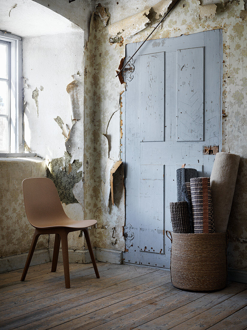 Wooden chair and rolled rugs in room with peeling wallpaper