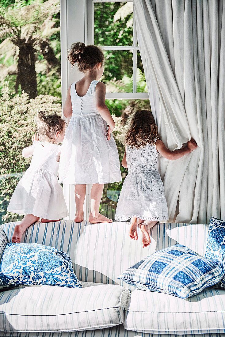 Three girls in white summer dresses climb around the window on a sofa