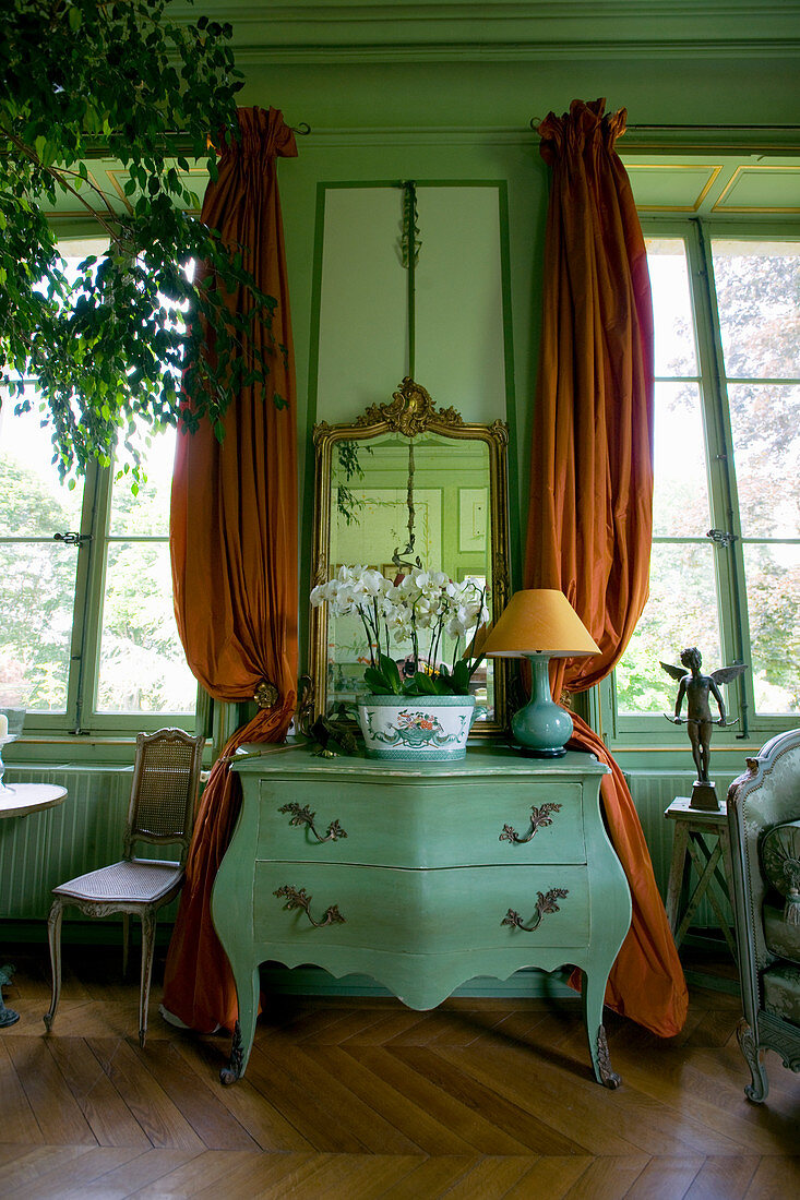 Zwischen Fenstern und Vorhängen platzierte antike Kommode in grünem französischen Salon