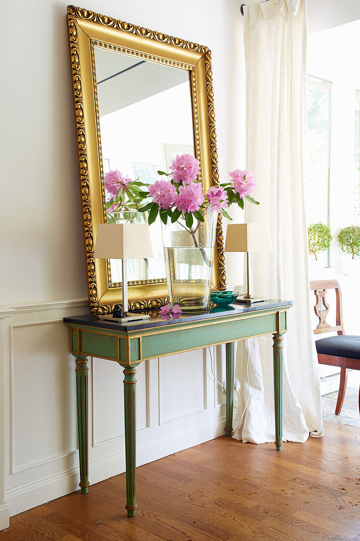 Gilt-framed mirror, vase of peonies and table lamps on antique console table