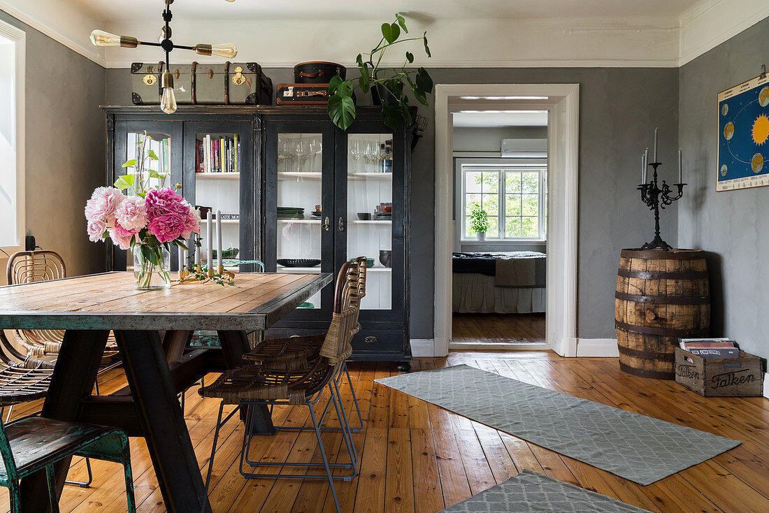 Dining table with rustic wooden top and vintage chairs in front of glass-fronted cabinet and old wooden barrel in corner of room