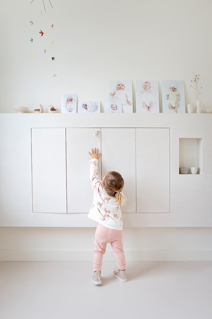Little girl standing in front of floating cupboards with baby photos on top