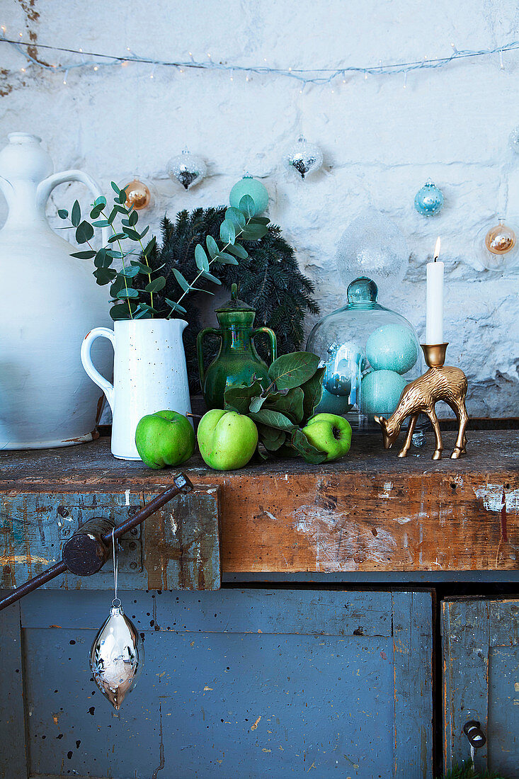 Krug mit Eukalyptuszweig, grüne Äpfel, Glasbehälter und Kerzenhalter auf altem Schrank