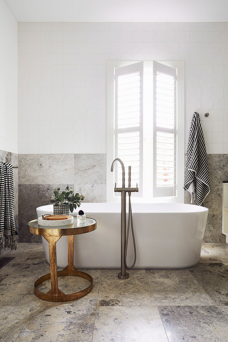 Freistehende Badewanne und goldener Beistelltisch vorm Fenster