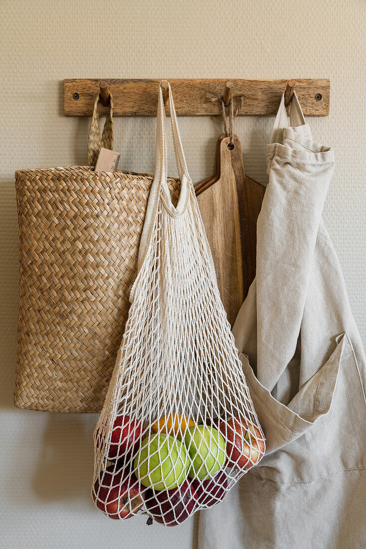 Net shopping bag of apples, raffia basket, chopping boards and apron hanging from row of hooks