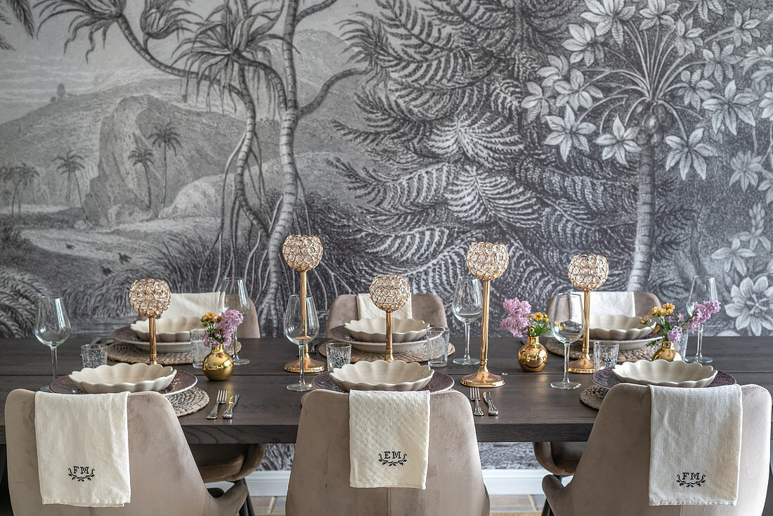 Festively set dining table with upholstered chairs in front of monochrome mural wallpaper
