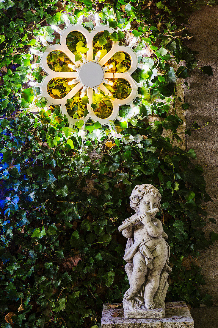Cherub playing flute below illuminated wreath on wall in garden