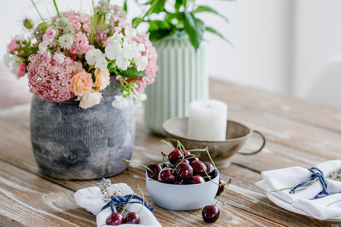 Vase of summer flowers, small bowl of cherries and candle on wooden table