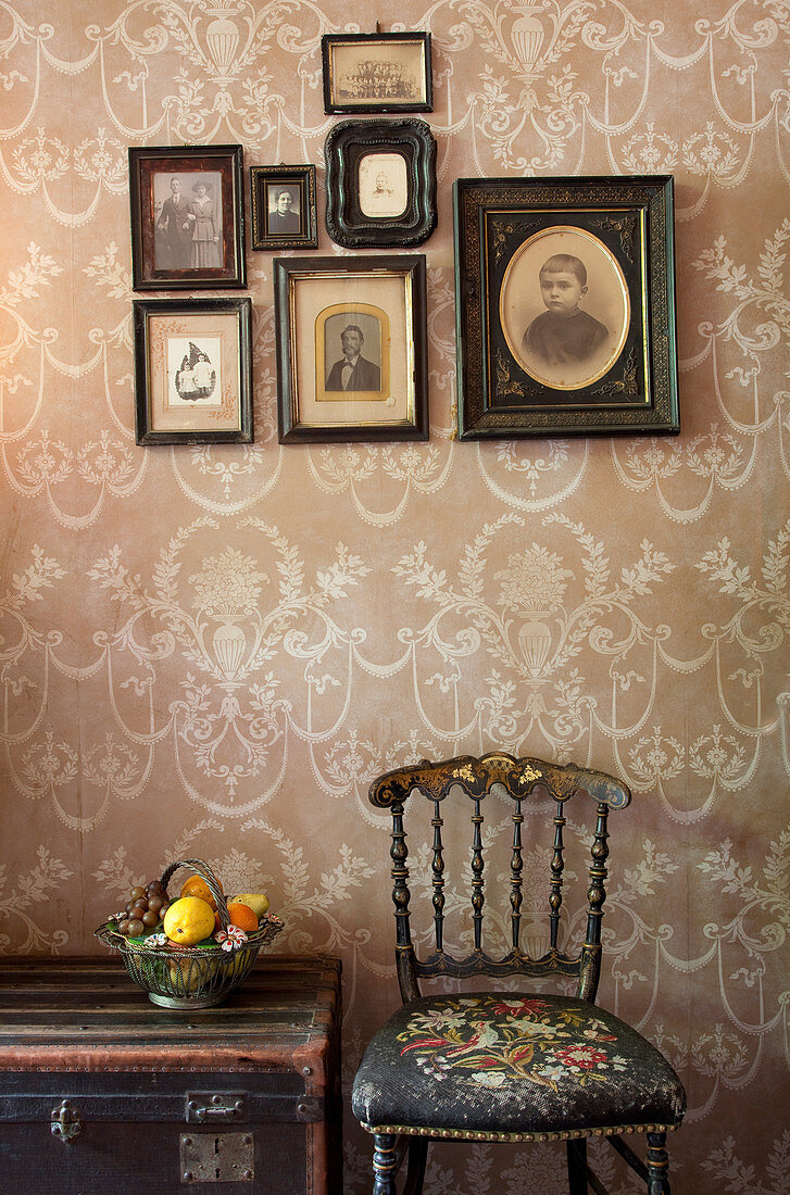 Antiker Stuhl und Truhe, darüber gerahmte Fotos an Wand mit Ornament Tapete
