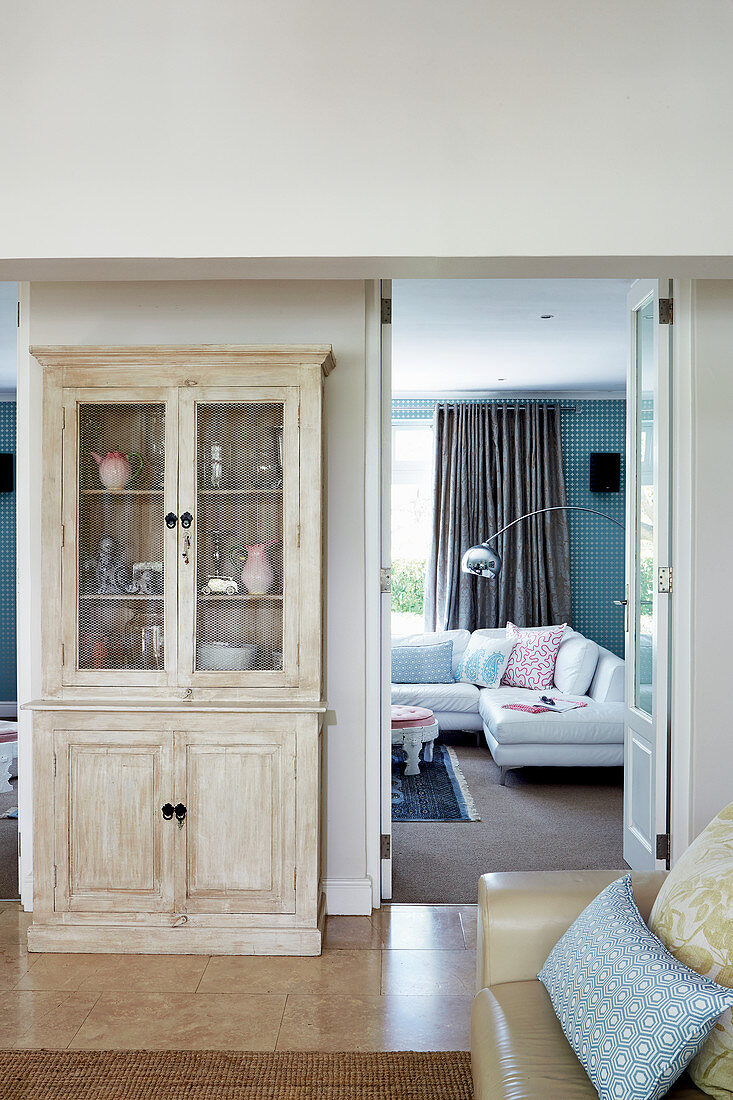 Simple glass-fronted cabinet made of pale wood next to door leading into colourful living room