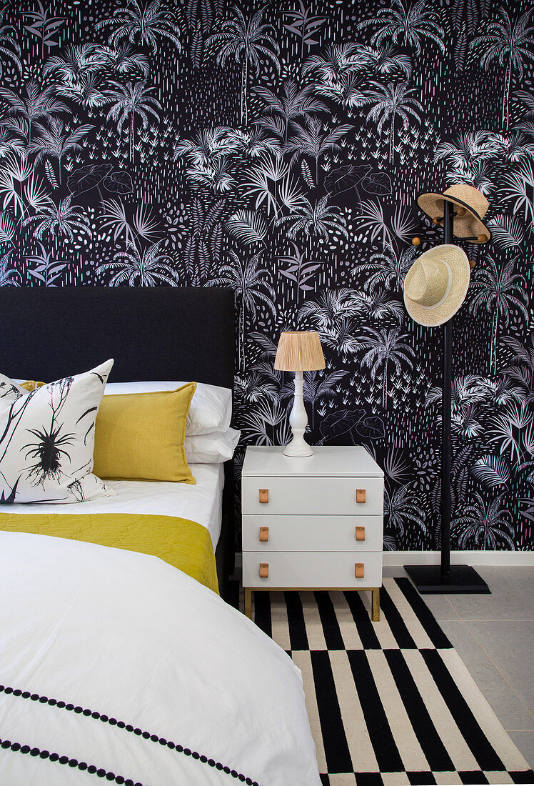 Bed and bedside cabinet against black wallpaper with plant motif in bedroom