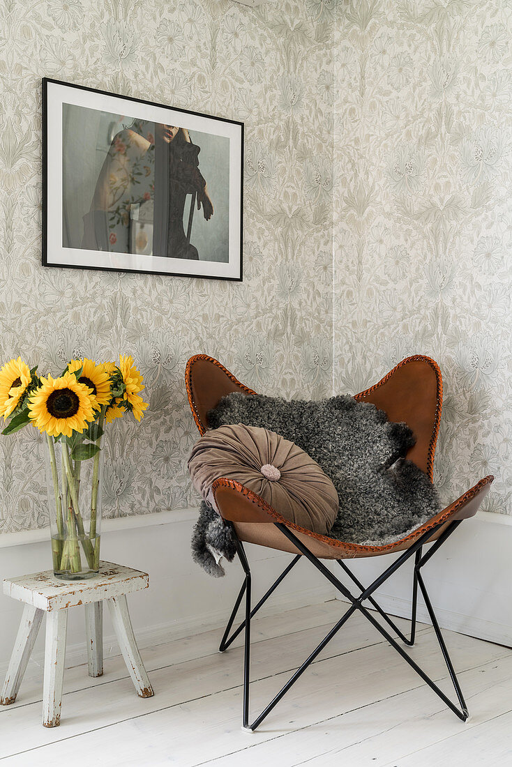 Fur blanket and velvet cushion on leather armchair in corner next to vase of sunflowers