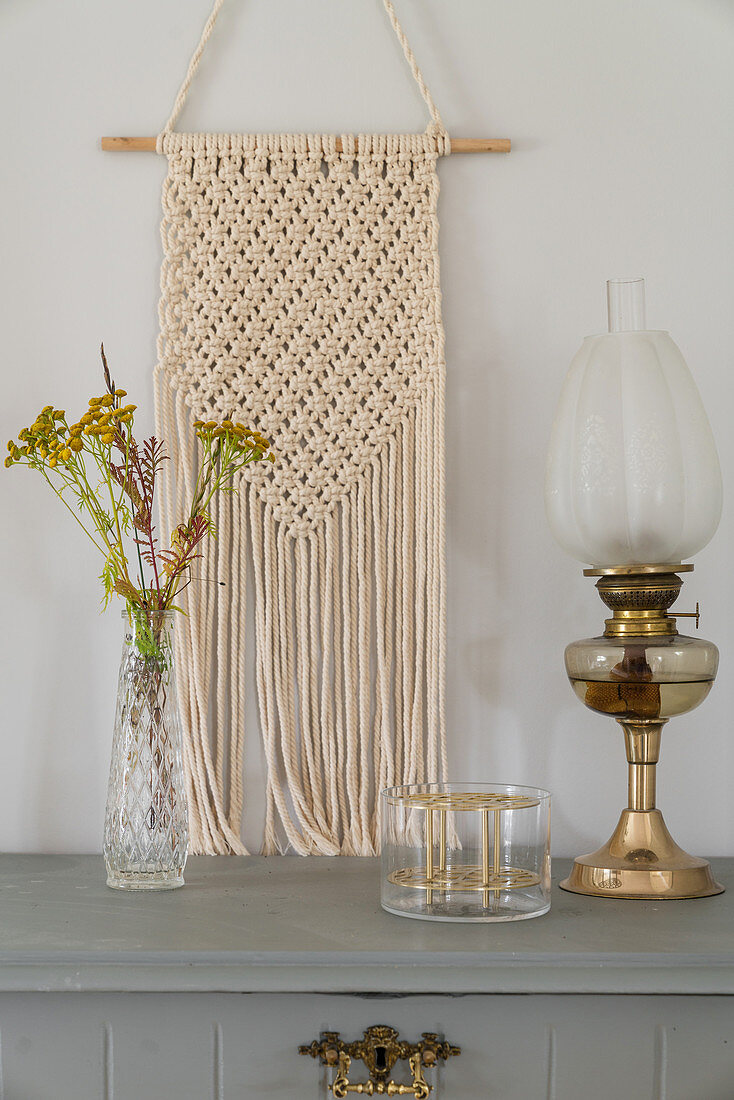 Macrame on pale grey wall above vase and oil lamp on grey cabinet