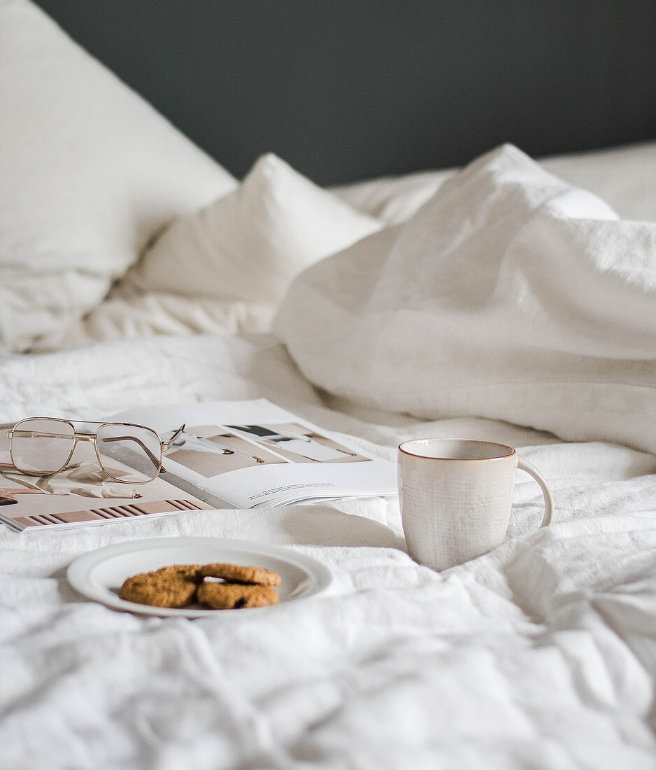 Plate of biscuits, mug, book and glasses on bed with white bed linen