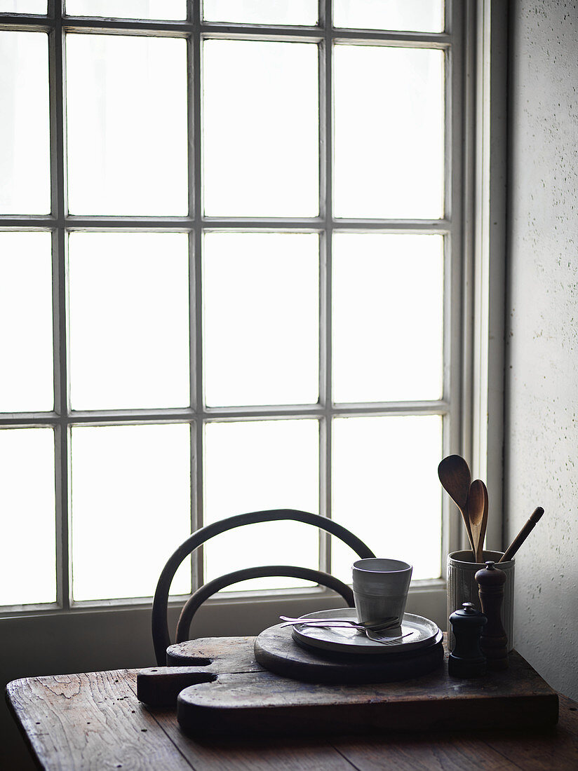 Schneidebrett mit Küchenutensilien und Geschirr auf Holztisch vor Fenster