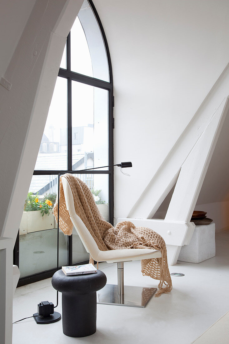 Blanket on modern easy chair and stool next to arched window