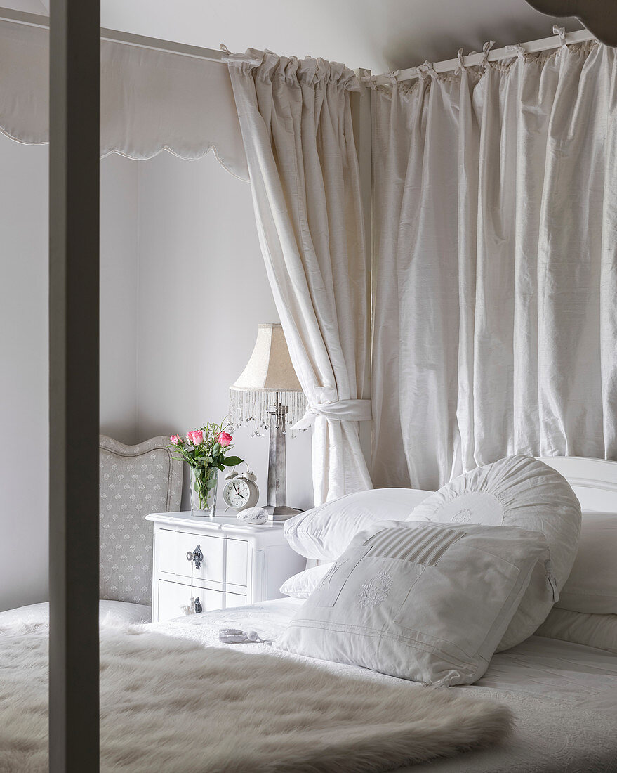 White cushions and fur blanket on four postered bed with cut roses and lamp at bedside