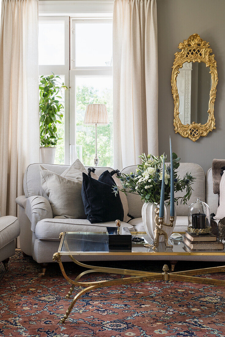 Antique glass table, upholstered sofa and gilt-framed mirror next to window in living room