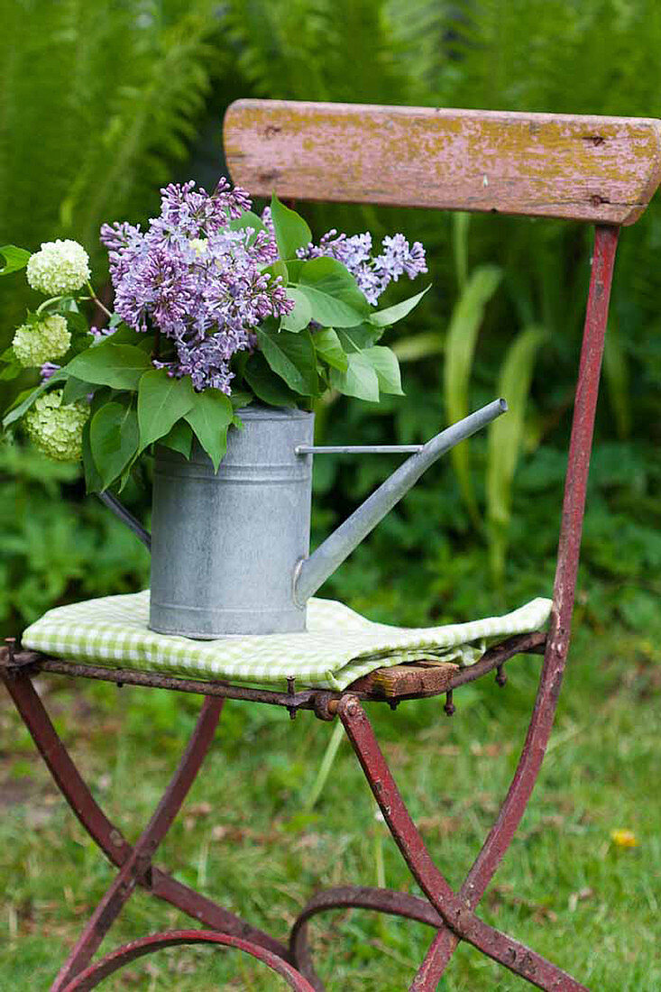 Bouquet of lilac and viburnum in zinc watering can on chair in garden