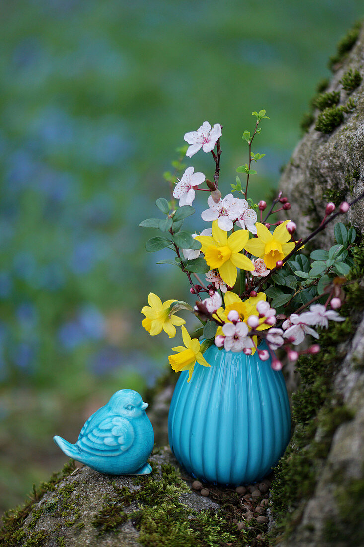 Spring bouquet of daffodils, blood plum, and boxwood in a turquoise vase next to a ceramic bluebird decoration