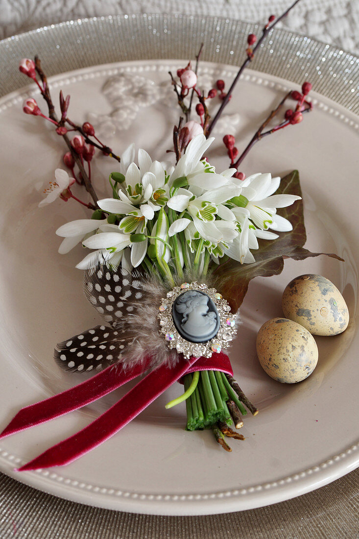 Posy of snowdrops, sprigs of purple-leaf plum tree blossom, feathers and ivy leaf decorating plate for Easter meal