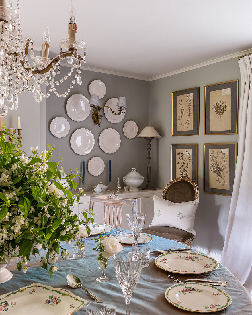 Gedeckter runder Tisch unter französischem Kronleuchter, Teller und botanische Drucke an den Wänden