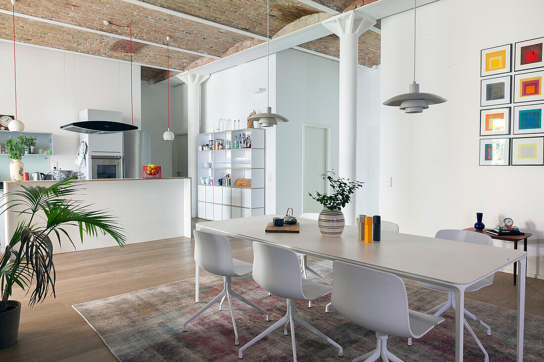 Weißer Esstisch mit Stühlen, darüber Pendelleuchte, im Hintergrund Küche in offenem Wohnraum mit hoher Decke