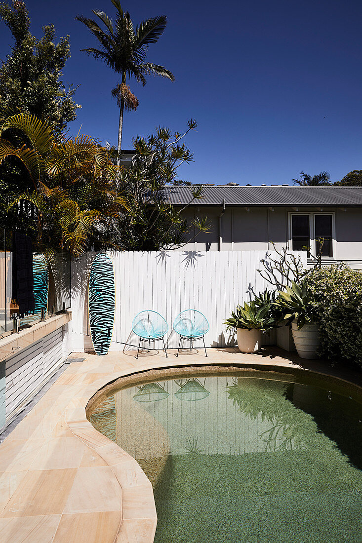 Swimming pool in garden with palm trees; surfboard leaning against the fence