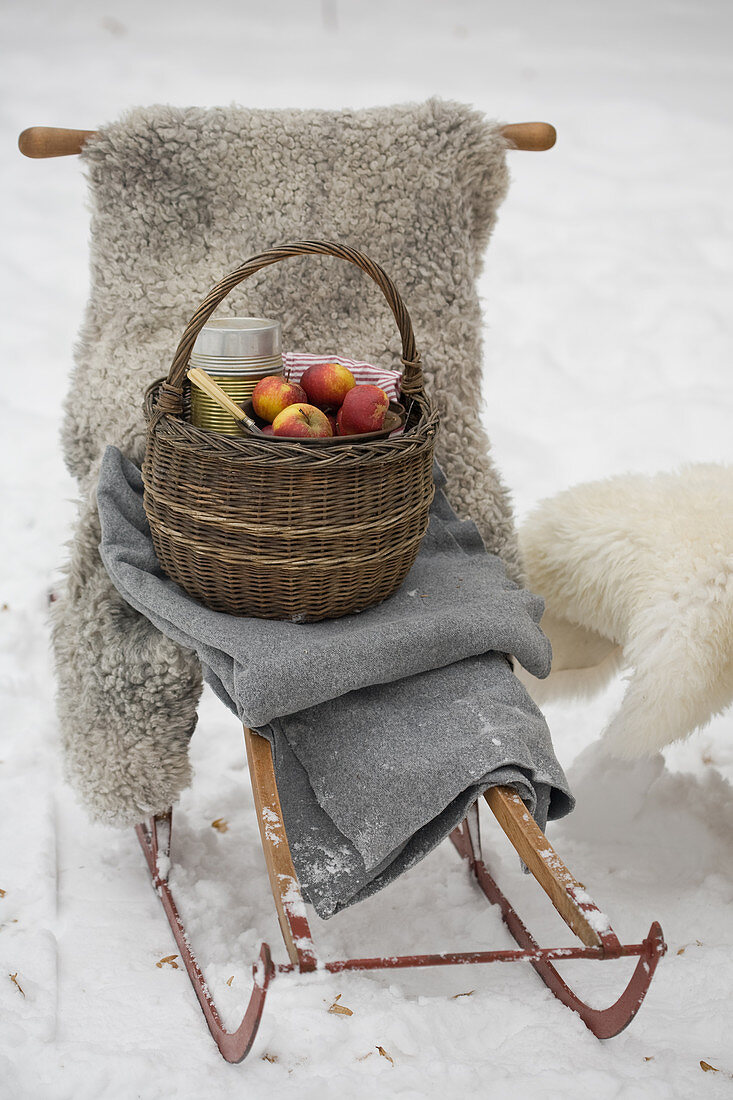 Korb mit Äpfeln und Thermosflasche auf Schlitten mit Schafsfell und Decke