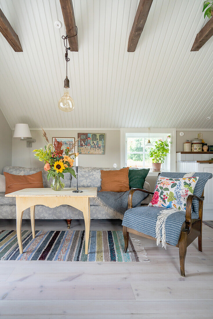 Sessel, Couchtisch mit Blumenstrauß und Sofa im Wohnraum mit weiß gestrichener Holzdecke