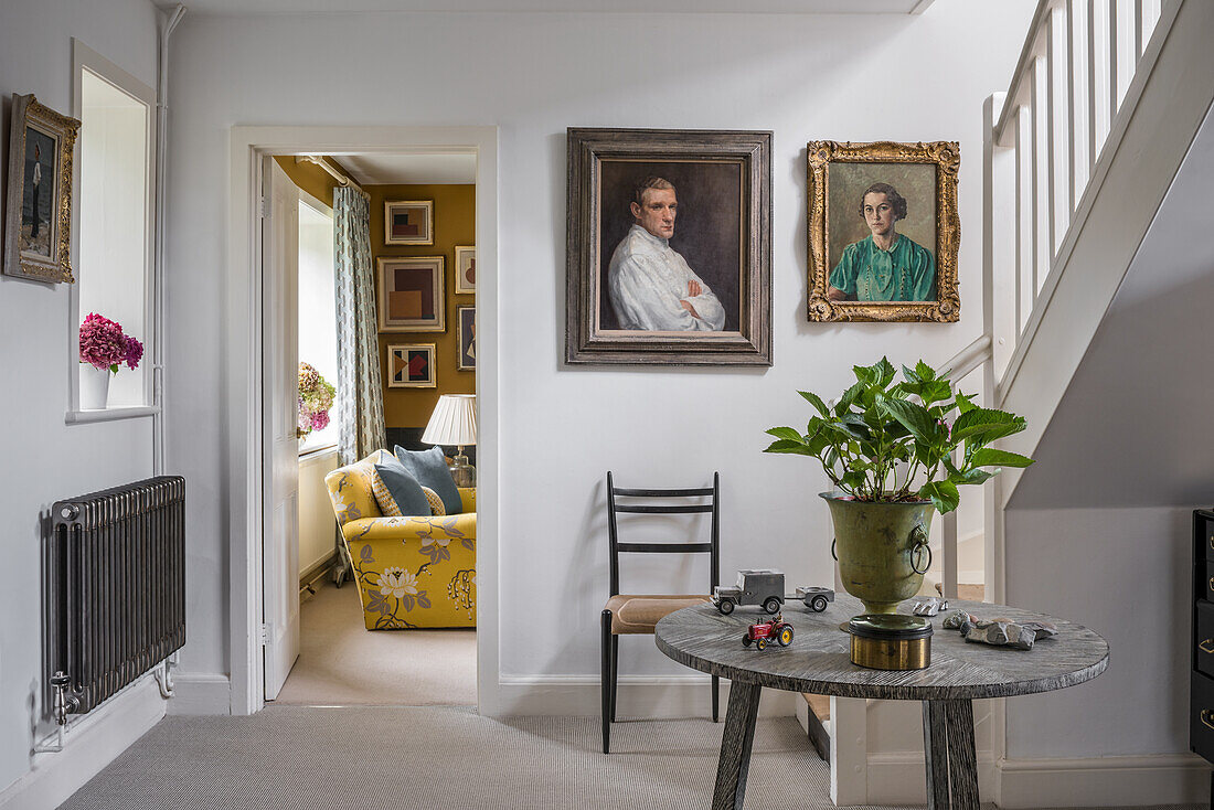 Houseplant on round marble table and portraits on wall in hallway with staircase