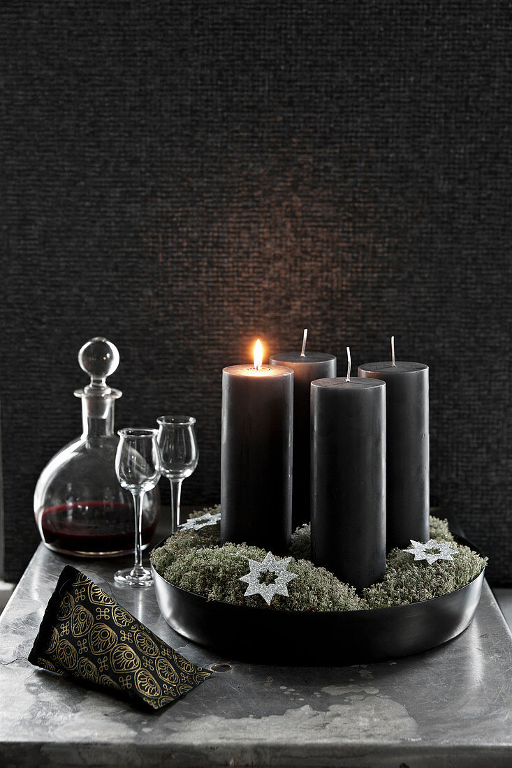 Black pillar candles, liqueur glasses and carafe in front of black wall